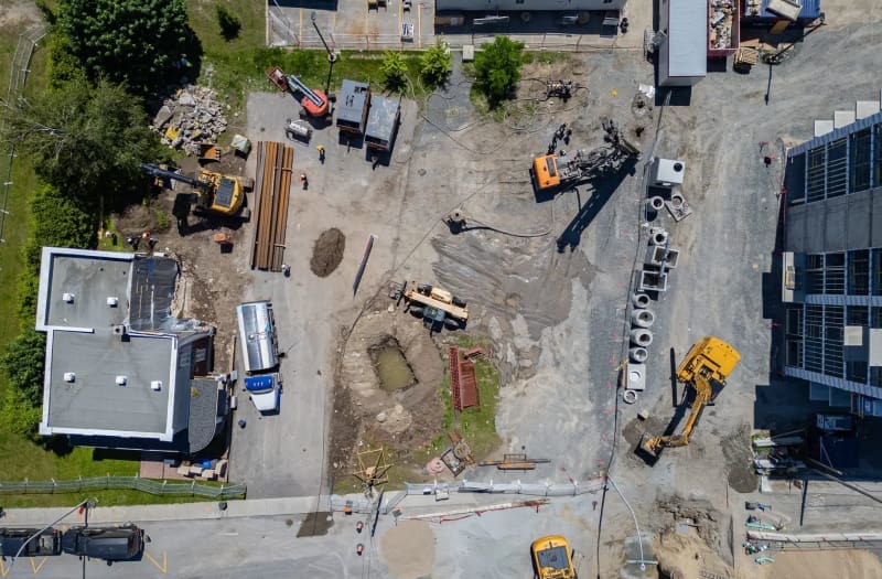 Vue aérienne d'un chantier de construction où des engins, du matériel, des matériaux et des ouvriers sont dispersés autour de bâtiments inachevés, de tas de terre et de tuyaux en béton, sur une zone partiellement pavée.