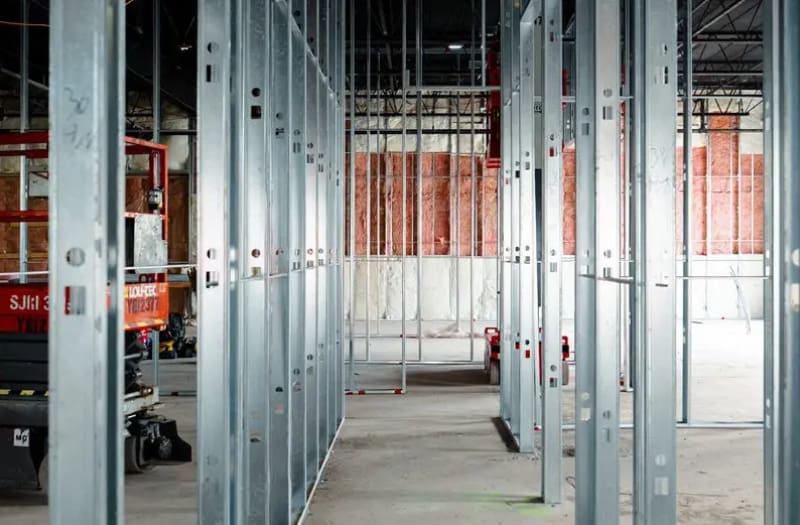 Des montants métalliques sont installés dans un bâtiment en cours de construction ; on aperçoit l'isolation en arrière-plan et du matériel de chantier sur le côté gauche de la scène.