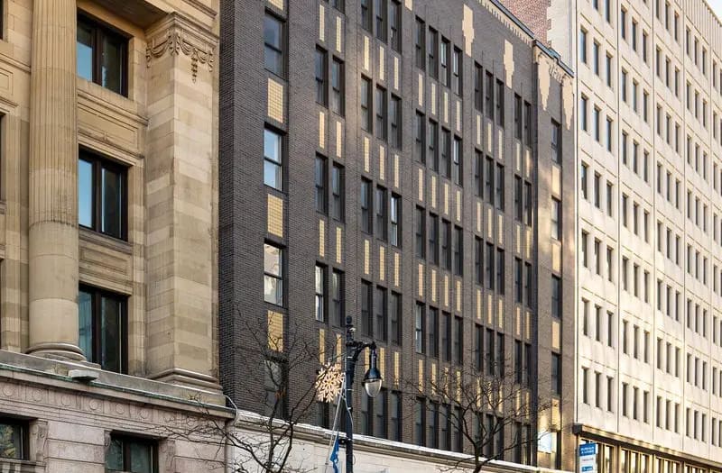 Une rangée d'immeubles urbains de plusieurs étages, aux styles architecturaux variés, avec notamment des façades en brique et en pierre, des fenêtres et des lampadaires ornés de motifs en forme de flocons de neige. Des arbres dépouillés bordent le trottoir.