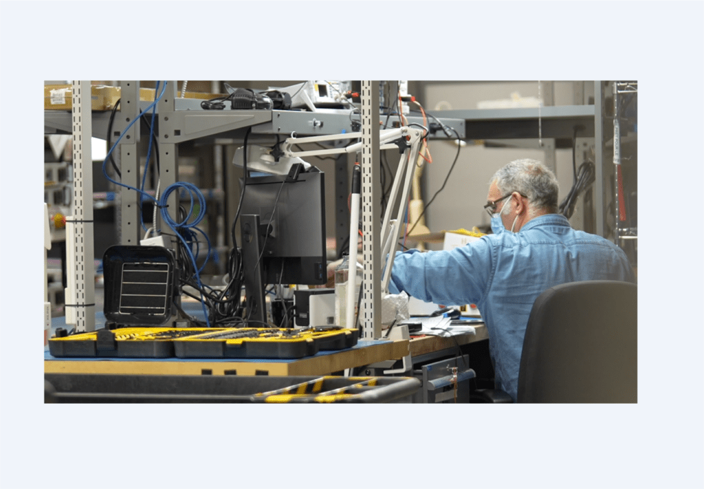 Un homme portant un masque et une chemise bleue travaille à un bureau dans un laboratoire d'électronique, entouré d'outils, de câbles et d'équipements. Des boîtes à outils jaunes sont visibles sur la table au premier plan.